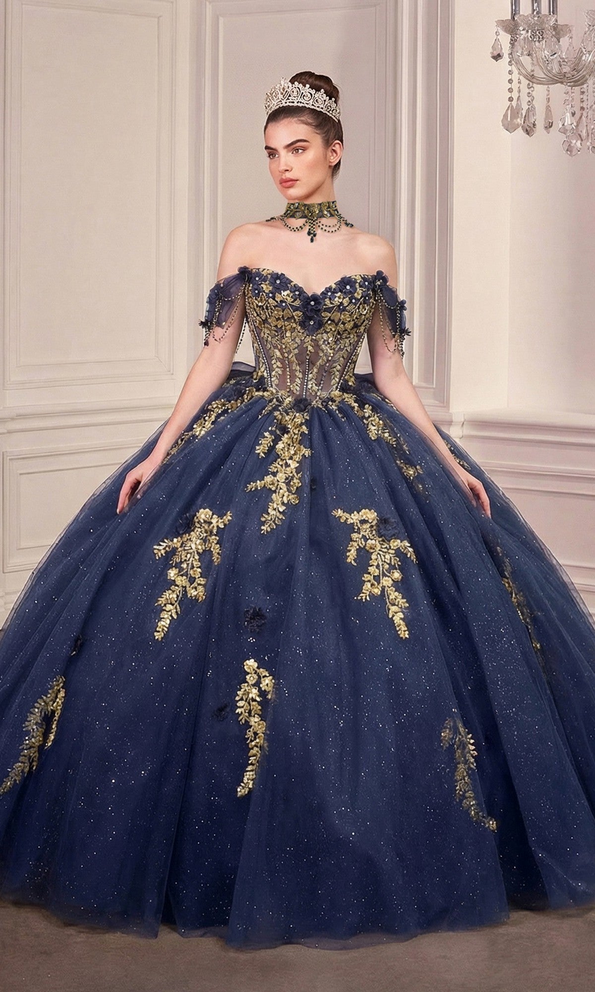 A woman in the Quinceanera Dress 15728 By Ladivine—navy blue and gold with off-the-shoulder sleeves, jeweled crown, and choker—stands in an elegant, light room with decorative panels and a chandelier.